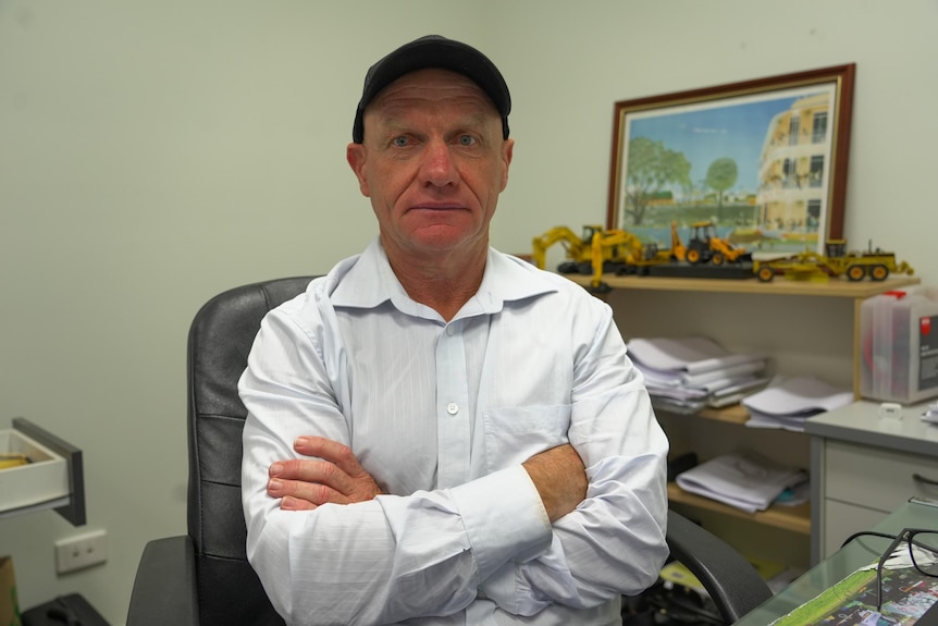 A man wearing a cap and a collared shirt sits at a desk with his arms crossed over his chest.