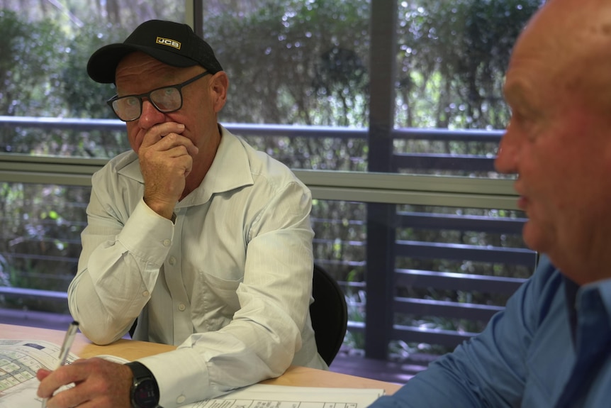 A man wearing a cap, glasses and a collared shirt sits at a table with one hand over his mouth.