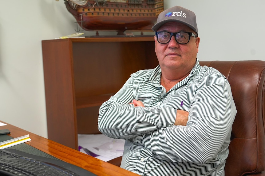 A man wearing glasses, a striped shirt and a cap sits at a desk.