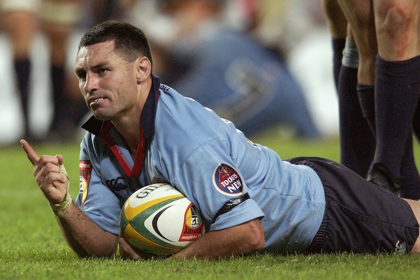 A male rugby player laying on a field on his front, holding a football in one hand and pointing a finger with his other hand.